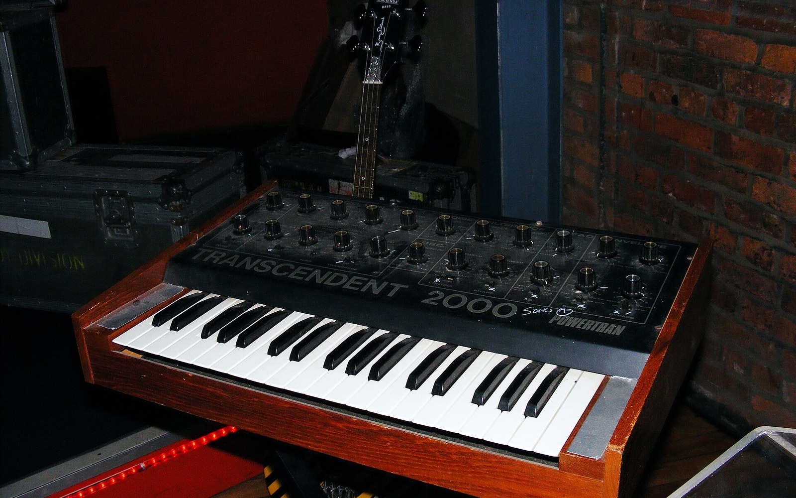 A Powertran Transcendent 2000 synthesizer with wooden case sits near flight cases and brick wall